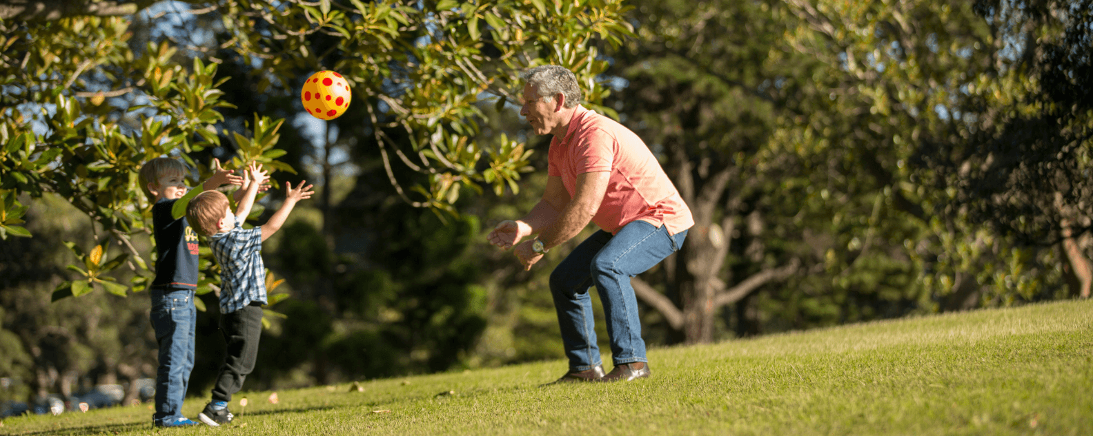 Two young children and an older male carer throw an orange ball under a fig tree.