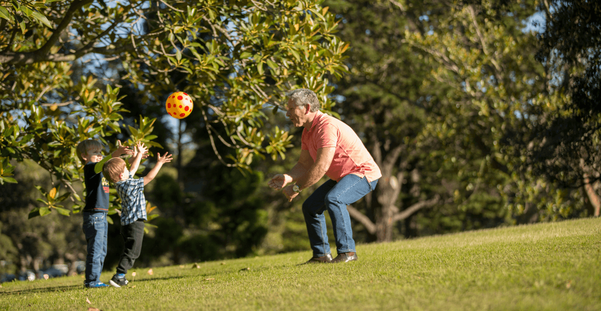 Two children throw a ball with an older man in the park under a tree