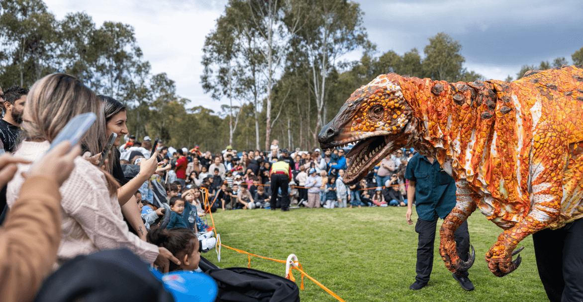 A life-sized orange dinosaur meets a large crowd of people outdoors in Lizard Log.