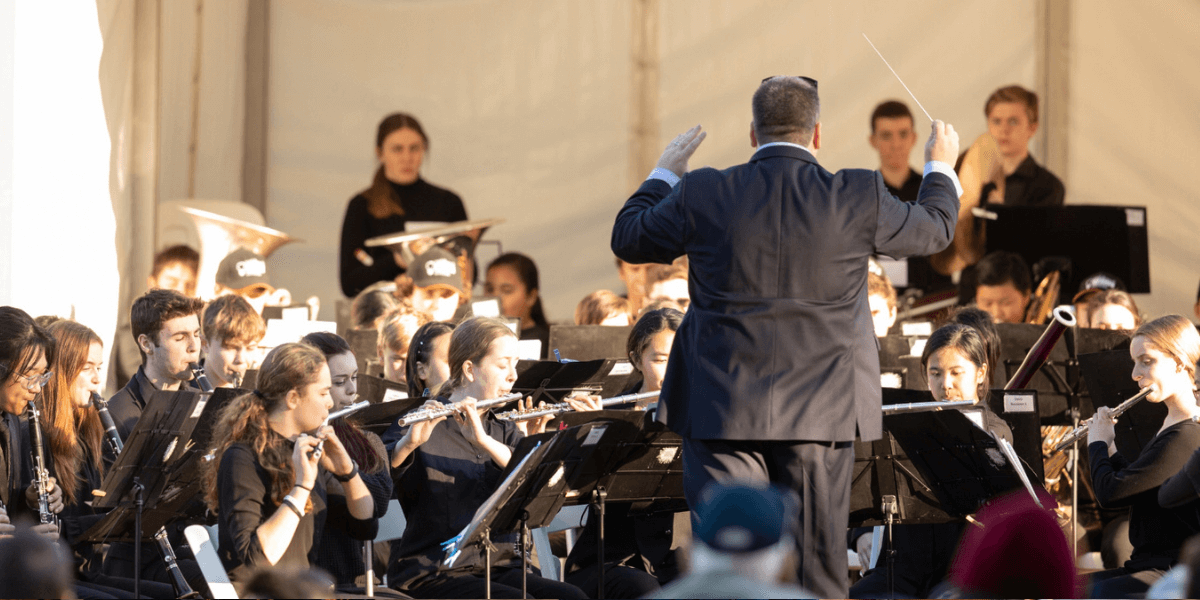 A conductor and orchestra perform outdoors in a marquee.