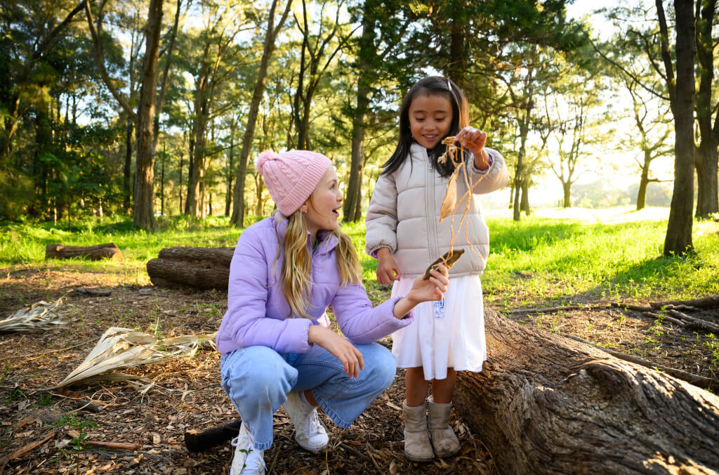 A young girl proudly displaying a nature-inspired craft to a smiling facilitator in a peaceful forest setting, surrounded by sunlight filtering through the trees.