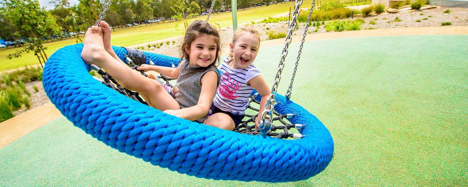 Two young girls happily swing together at Domain Creek Playground, enjoying a sunny day filled with laughter and fun.