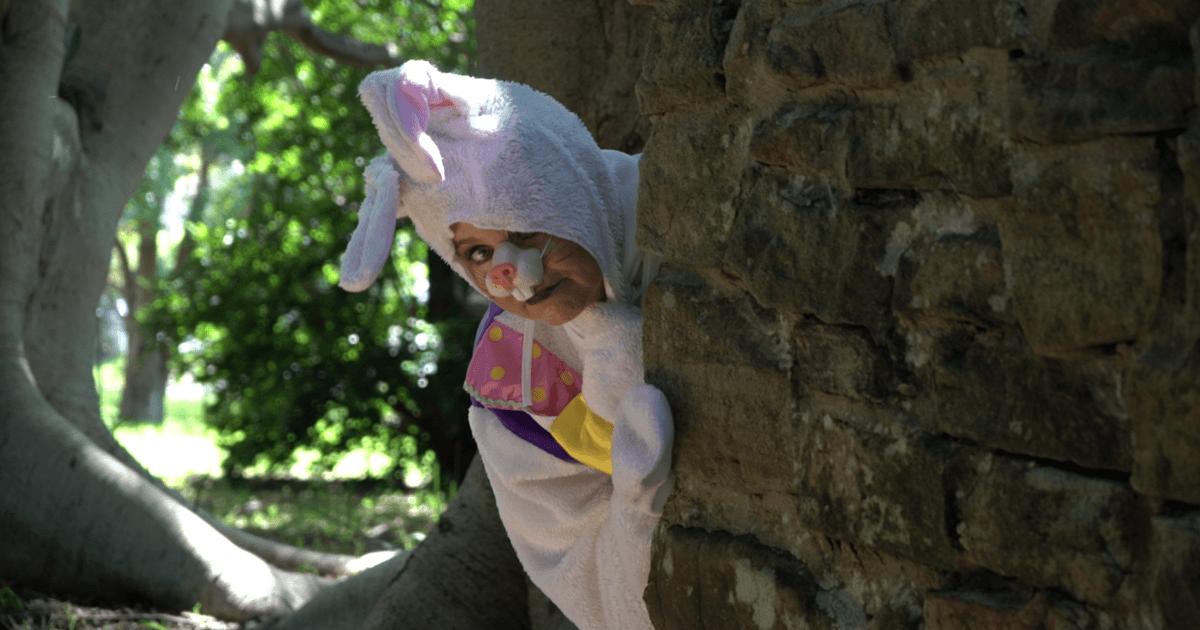 A person dressed in a playful Easter bunny costume with a purple, yellow, and pink vest peeks around a stone wall, surrounded by green trees.