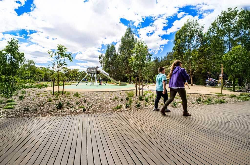  Two people stroll along a wooden walkway beside Domain Creek Playground, enjoying a sunny day outdoors.