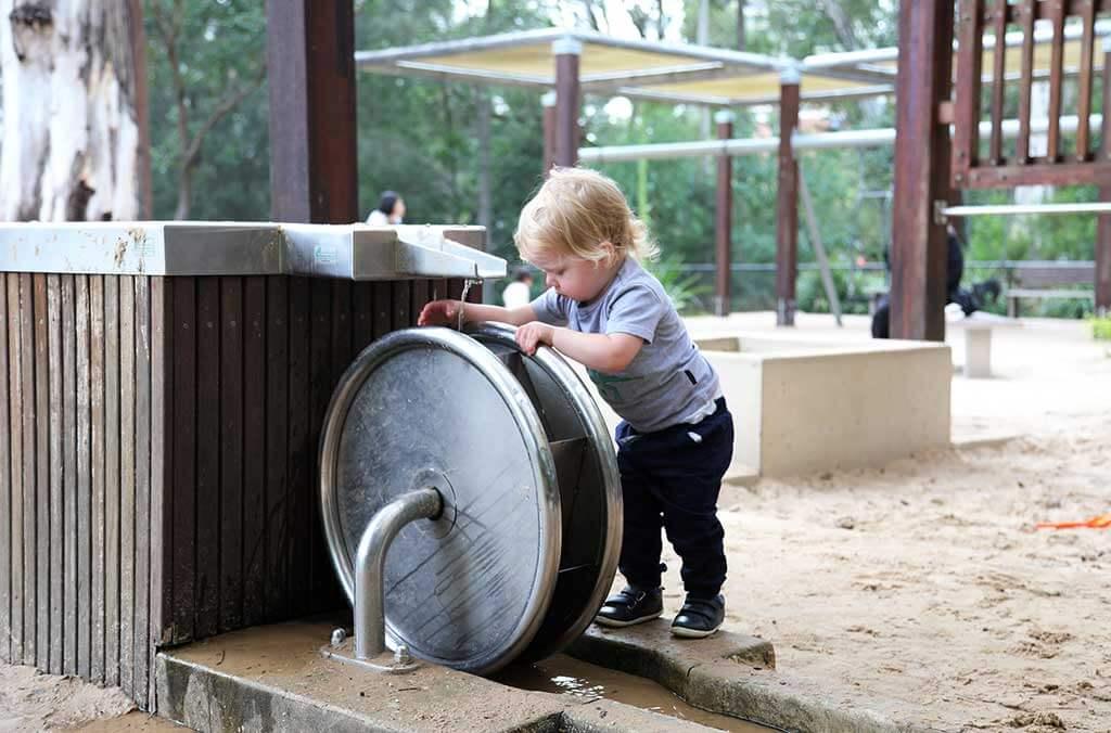 A young child plays with a big metal wheel at Paperbark Playground, surrounded by nature and fun.