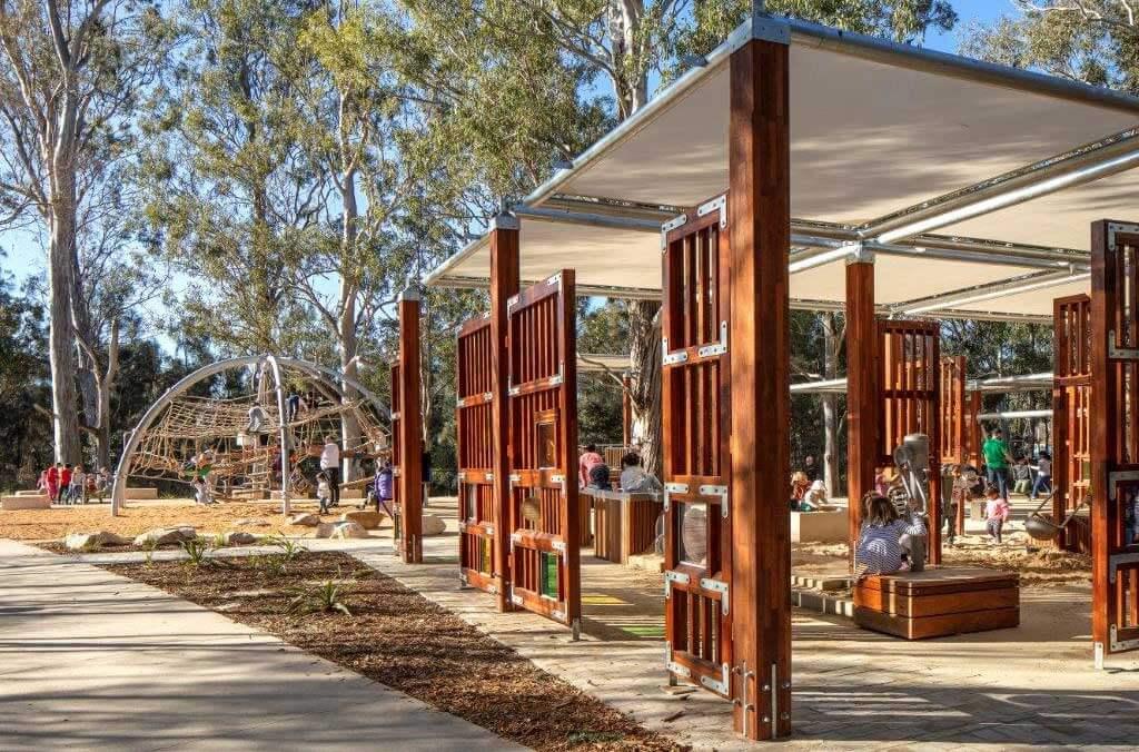 People enjoy the shade of a wooden pavilion at Paperbark Playground, sitting on benches and having a good time together.